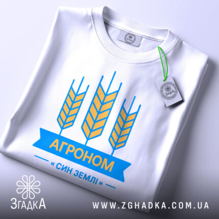 Футболка агроном син землі, біла, складена на поверхні, зображена з верхнього ракурсу, фон світлий, принт з жовтими колосками та синьою стрічкою з написом "АГРОНОМ", на ярлику зелена стрічка.
