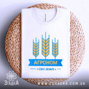 Футболка агроном син землі, біла, складена на плетеній кошику, зображена з верхнього ракурсу, фон світлий, принт з жовтими колосками та синьою стрічкою з написом "АГРОНОМ".