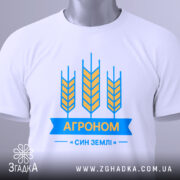 Футболка агроном син землі, біла, складена на поверхні, зображена з верхнього ракурсу, фон світлий, принт з жовтими колосками та синьою стрічкою з написом "АГРОНОМ", на ярлику зелена стрічка.