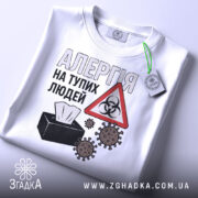 Футболка алергія люди. Біла футболка, складена на поверхні, з видимим коміром. На фоні світло-сірий градієнт. Принт з текстом 'АЛЕРГІЯ НА ТУПИХ ЛЮДЕЙ', символ небезпеки та малюнки мікробів, а також етикетка з зеленим шнурком.