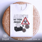 Футболка алергія люди. Біла футболка, складена на плетеній основі. На фоні світло-сірий градієнт. Принт з текстом 'АЛЕРГІЯ НА ТУПИХ ЛЮДЕЙ', символ небезпеки та малюнки мікробів.