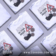 Футболка алергія люди. Біла футболка з коротким рукавом, зображена в ракурсі спереду. На фоні світло-сірий градієнт. Принт містить текст 'АЛЕРГІЯ НА ТУПИХ ЛЮДЕЙ', символ небезпеки та малюнки мікробів.