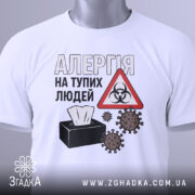 Футболка алергія люди. Біла футболка, складена на поверхні, з видимим коміром. На фоні світло-сірий градієнт. Принт з текстом 'АЛЕРГІЯ НА ТУПИХ ЛЮДЕЙ', символ небезпеки та малюнки мікробів, а також етикетка з зеленим шнурком.