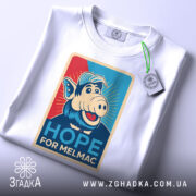 Футболка Альф білого кольору, складена на столі, ракурс зверху, фон світло-сірий, принт з персонажем свині в кепці, текст "HOPE FOR MELMAC" з елементами графіки.