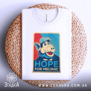 Футболка Альф білого кольору, складена на плетеній кошику, ракурс зверху, фон світло-сірий, принт з персонажем свині в кепці, текст "HOPE FOR MELMAC" на яскравому фоні.