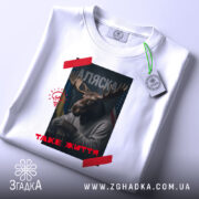 Футболка Аляска таке життя, складена біла футболка з принтом, зображення оленя з рогами, на фоні прапорів України та США, текст 'ТАKE ЖИТТЯ' червоним кольором, етикетка з зеленим шнурком.