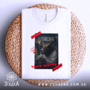 Футболка Аляска таке життя, складена біла футболка з принтом, зображення оленя з рогами, на фоні прапорів України та США, текст 'ТАKE ЖИТТЯ' червоним кольором, розташована на плетеній основі.