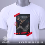Футболка Аляска таке життя, складена біла футболка з принтом, зображення оленя з рогами, на фоні прапорів України та США, текст 'ТАKE ЖИТТЯ' червоним кольором, етикетка з зеленим шнурком.
