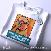 Футболка Алло заберіть мене. Біла футболка, складена на поверхні, з принтом мавпи, що тримає телефон. Текст під принтом синій, на фоні оранжевих відтінків. На комірці видно етикетку.
