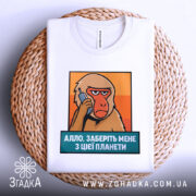 Футболка Алло заберіть мене. Біла футболка, складена на плетеній основі, з принтом мавпи, що тримає телефон. Текст під принтом синій, фон оранжевий. Видно етикетку на комірці.