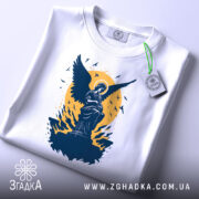 Футболка ангел України, біла, складена на столі, фон світло-сірий, принт з ангелом на фоні жовтого кола, з етикеткою зеленого кольору.