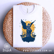 Футболка ангел України, біла, складена на кошику з плетеного матеріалу, фон світло-сірий, принт з ангелом на фоні жовтого кола та птахів.