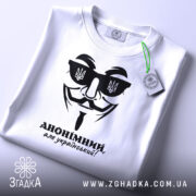 Футболка анонімний український, біла, складена на плоскій поверхні, фон світлий, принт чорного кольору з зображенням маски та написом 'АНОНІМНИЙ, але український!', на комірці видно етикетку.