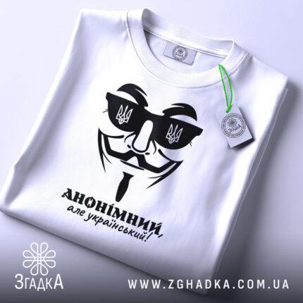 Футболка анонімний український, біла, складена на плоскій поверхні, фон світлий, принт чорного кольору з зображенням маски та написом 'АНОНІМНИЙ, але український!', на комірці видно етикетку.