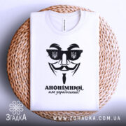 Футболка анонімний український, біла, складена на плоскій поверхні, фон світлий, принт чорного кольору з зображенням маски та написом 'АНОНІМНИЙ, але український!', на фоні плетена кошик.