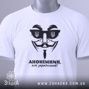Футболка анонімний український, біла, складена на плоскій поверхні, фон світлий, принт чорного кольору з зображенням маски та написом 'АНОНІМНИЙ, але український!', на комірці видно етикетку.