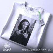 Футболка античний стріт, біла, складена на столі, фон світло-сірий, принт з чорно-білим зображенням статуї в капюшоні, яка показує жест, з етикеткою на шнурку.