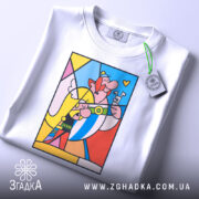 Футболка арт герої білого кольору, зображена зверху на плоскій поверхні, фон світло-сірий. Принт яскравий, з кольоровими персонажами, що посміхаються, з елементами жовтого, червоного та блакитного кольорів, з прикріпленою етикеткою.