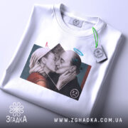 Футболка арт кохання, біла, складена на столі, фон світло-сірий, принт з двома людьми, що цілуються, з елементами графіки, бірка з зеленим шнурком.