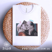 Футболка арт кохання, біла, складена на кошику, фон світло-сірий, принт з двома людьми, що цілуються, з елементами графіки, вид зверху.