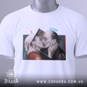 Футболка арт кохання, біла, складена на столі, фон світло-сірий, принт з двома людьми, що цілуються, з елементами графіки, бірка з зеленим шнурком.