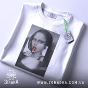 Футболка арт лоліпоп, біла, складена на поверхні, фон світло-сірий, принт з жінкою з червоним льодяником, на комірці видна етикетка.