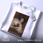 Футболка арт нестримні веселощі, біла, зображена зверху, на складеній футболці, фон світло-блакитний, принт з зображенням дівчини з котом, з етикеткою на рукаві.