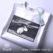 Футболка арт заєць білого кольору, складена на поверхні, ракурс зверху, фон світло-сірий, принт з зображенням зайця в стилі лінійного малюнка, прикріплена етикетка з зеленим шнурком.