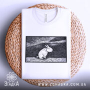 Футболка арт заєць білого кольору, складена на кошику, ракурс зверху, фон світло-сірий, принт з зображенням зайця в стилі лінійного малюнка на чорному фоні.