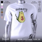 Футболка авокадо котик, білого кольору, зображена на манекені з правого боку, фон світло-сірий, принт з авокадо-котом, що тримає червону кульку, текст 'AVOGATO ¡hola!' над малюнком.