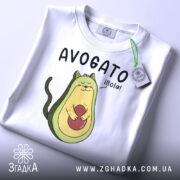 Футболка авокадо котик, білого кольору, складена на столі, фон світло-сірий, принт з авокадо-котом, що тримає червону кульку, текст 'AVOGATO ¡hola!' над малюнком, з етикеткою на зеленій стрічці.