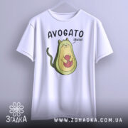 Футболка авокадо котик, білого кольору, підвішена на вішалці, фон світло-сірий, принт з авокадо-котом, що тримає червону кульку, текст 'AVOGATO ¡hola!' над малюнком.