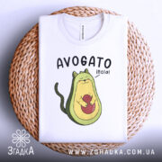 Футболка авокадо котик, білого кольору, складена на плетеній кошику, фон білий, принт з авокадо-котом, що тримає червону кульку, текст 'AVOGATO ¡hola!' над малюнком.