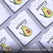Футболка авокадо котик, білого кольору, зображена на манекені з правого боку, фон світло-сірий, принт з авокадо-котом, що тримає червону кульку, текст 'AVOGATO ¡hola!' над малюнком.