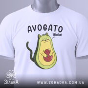 Футболка авокадо котик, білого кольору, складена на столі, фон світло-сірий, принт з авокадо-котом, що тримає червону кульку, текст 'AVOGATO ¡hola!' над малюнком, з етикеткою на зеленій стрічці.