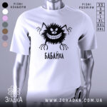 Футболка бабайка білого кольору, зображена з близького ракурсу на манекені, фон світло-сірий, принт чорного кольору з веселим персонажем та написом 'БАБАЙКА'.