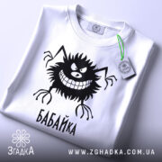 Футболка бабайка білого кольору, складена на поверхні, фон світло-сірий, принт чорного кольору з персонажем та написом 'БАБАЙКА', на комірці є етикетка з зеленим шнурком.