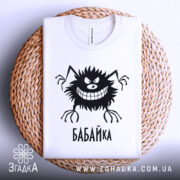 Футболка бабайка білого кольору, складена на плетеній кошику, фон світло-сірий, принт чорного кольору з персонажем та написом 'БАБАЙКА'.