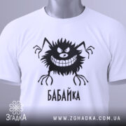 Футболка бабайка білого кольору, складена на поверхні, фон світло-сірий, принт чорного кольору з персонажем та написом 'БАБАЙКА', на комірці є етикетка з зеленим шнурком.