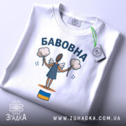 Футболка бабовна білого кольору, складена на поверхні, фон світло-сірий, принт з малюнком ангела з бавовняними кульками та написом 'БАБОВНА', на футболці є етикетка з зеленим шнурком.