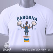 Футболка бабовна білого кольору, складена на поверхні, фон світло-сірий, принт з малюнком ангела з бавовняними кульками та написом 'БАБОВНА', на футболці є етикетка з зеленим шнурком.