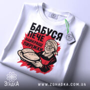 Футболка бабуся пече пиріжки, біла, зображена зверху, фон світло-сірий, принт з бабусею, що тримає пиріжки, з етикеткою на рукаві.