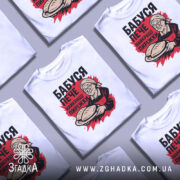 Футболка бабуся пече пиріжки, біла, зображена на манекені, фон світло-сірий, принт з бабусею, що тримає пиріжки, червоний фон за принтом.