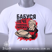 Футболка бабуся пече пиріжки, біла, зображена зверху, фон світло-сірий, принт з бабусею, що тримає пиріжки, з етикеткою на рукаві.