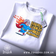 Футболка бабуся супергерой, біла, зображена на столі, фон світло-сірий, принт з бабусею-супергероєм у синьому костюмі, що біжить, з червоним плащем, напис 'Бабуся завжди прийде на допомогу', з етикеткою на комірці.