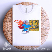 Футболка бабуся супергерой, біла, зображена на плетеному кошику, фон білий, принт з бабусею-супергероєм у синьому костюмі з червоним плащем, що біжить, з написом 'Бабуся завжди прийде на допомогу'.