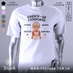 Футболка бабуся це суперсила, біла футболка з принтом, що зображує бабусю з окулярами, на фоні світло-сірого кольору, текст на футболці говорить 'Бабуся – це суперсила', оточений зірочками та сердечками.