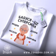 Футболка бабуся це суперсила, біла футболка, зображена зверху, на фоні світло-сірого кольору, принт з бабусею та текстом 'Бабуся – це суперсила', з зеленим ярликом, що звисає з коміра.