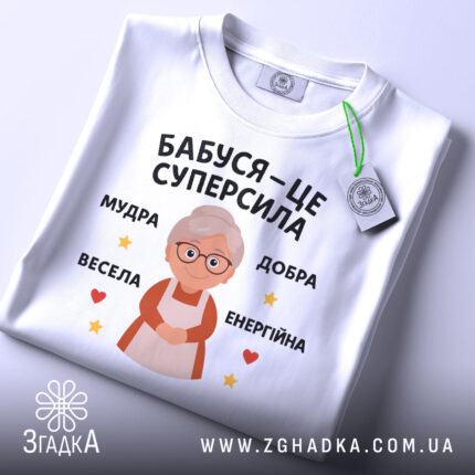 Футболка бабуся це суперсила, біла футболка, зображена зверху, на фоні світло-сірого кольору, принт з бабусею та текстом 'Бабуся – це суперсила', з зеленим ярликом, що звисає з коміра.