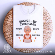 Футболка бабуся це суперсила, біла футболка, складена на плетеній основі, на фоні світло-сірого кольору, принт з бабусею та текстом 'Бабуся – це суперсила', оточений зірочками та сердечками.